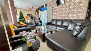 A modern living room with a black leather sofa, a glass coffee table, and a small side table. A Christmas tree and a vase with flowers are visible in the background. - Atreya Hotel Makassar (Makassar)