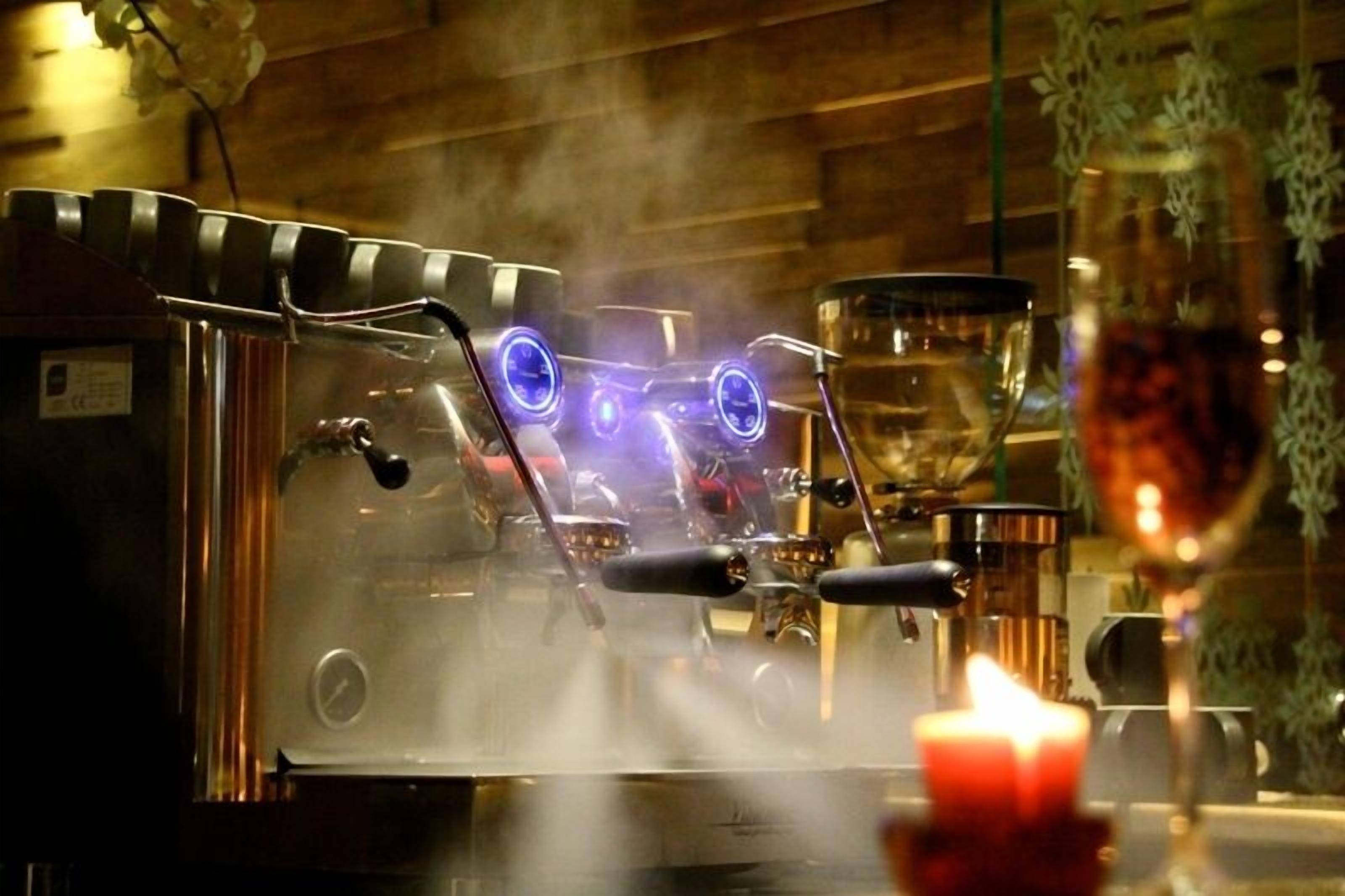 A coffee machine with steam, a glass of red wine, and a candle on a table.