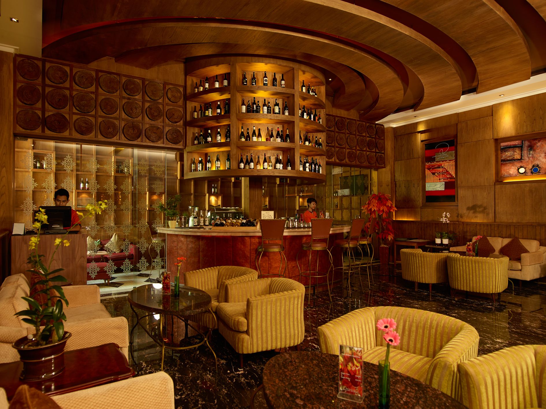 A bar area with multiple shelves of bottles, a counter, and bar stools. There are sofas and a small table with a vase of flowers.