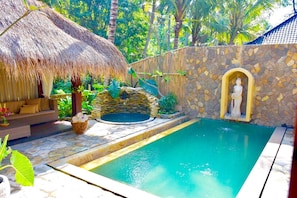 A swimming pool with a stone wall and a statue, surrounded by tropical plants and a thatched-roof structure. - Atman Villas (Ubud)