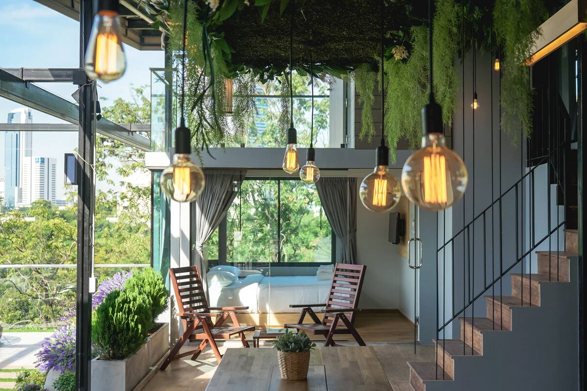 Una habitación moderna con suelo de madera, una cama con ropa blanca, dos sillas de madera, una mesita con una planta y una escalera con barandilla negra.