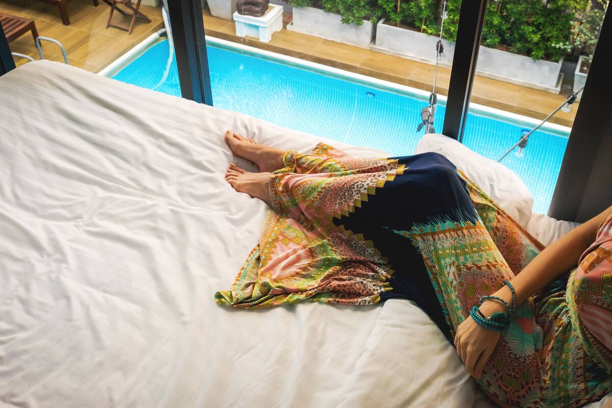 Una persona acostada en una cama con vista a una piscina a través de grandes ventanales.