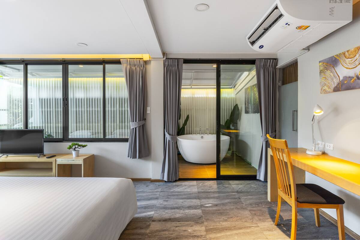 Una habitación de hotel moderna con cama, escritorio, televisor, baño con bañera y un amplio ventanal con cortinas.