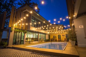 A modern building with a swimming pool, outdoor seating, and string lights.
