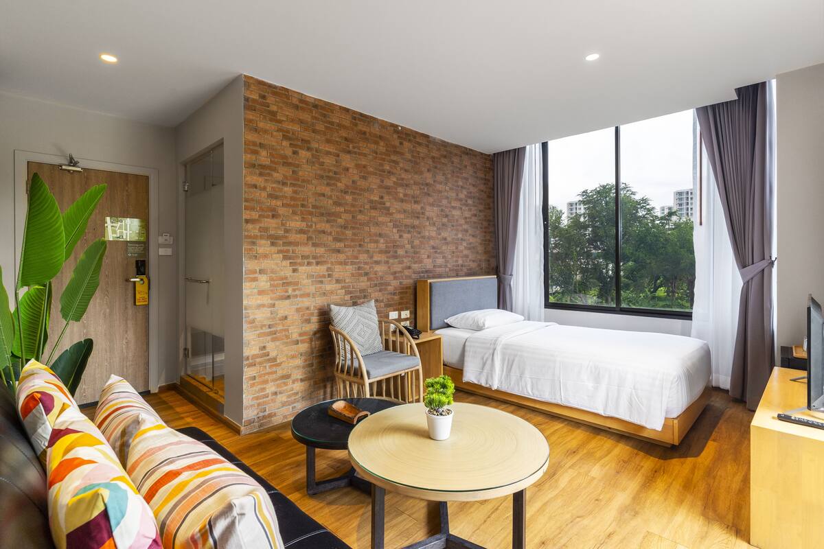 Una habitación de hotel moderna con una pared de ladrillo decorativa, una cama grande, una pequeña mesa redonda con una planta, un sofá con cojines coloridos y vistas al exterior a través de amplias ventanas.