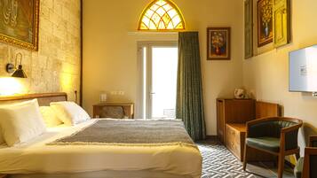 Quarto de hotel com cama, mesa de cabeceira de madeira, escrivaninha, cadeira, televisão e janela com detalhe em vitral.