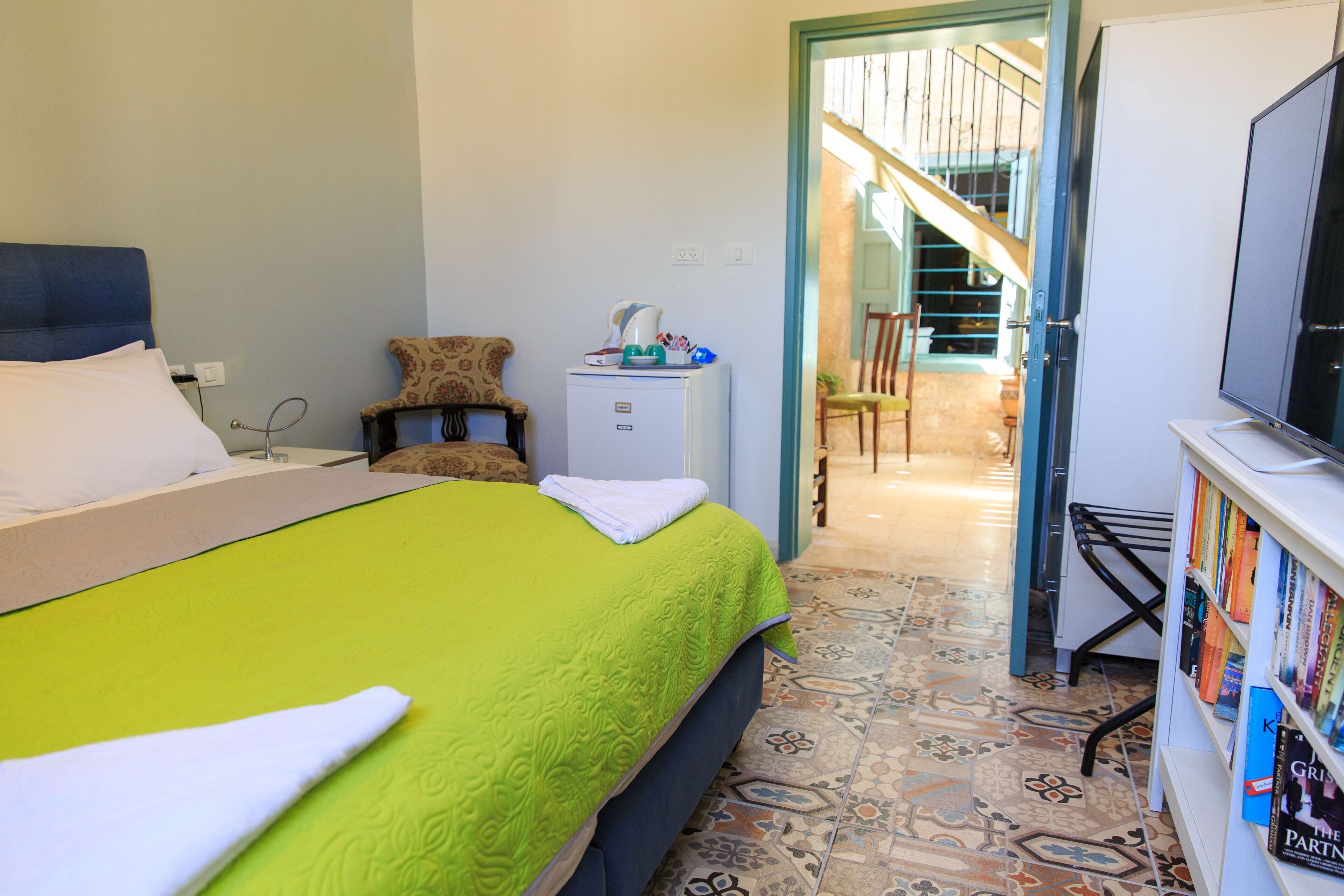 A hotel room with a bed, a small desk with a lamp, a chair, a bookshelf, and a staircase leading to another area.