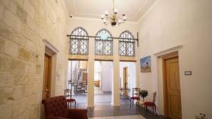 A spacious hallway with a high ceiling, stone walls, and a chandelier. There are multiple doors, a staircase, and a dining area with chairs and a table. - Michel House (Nazareth)