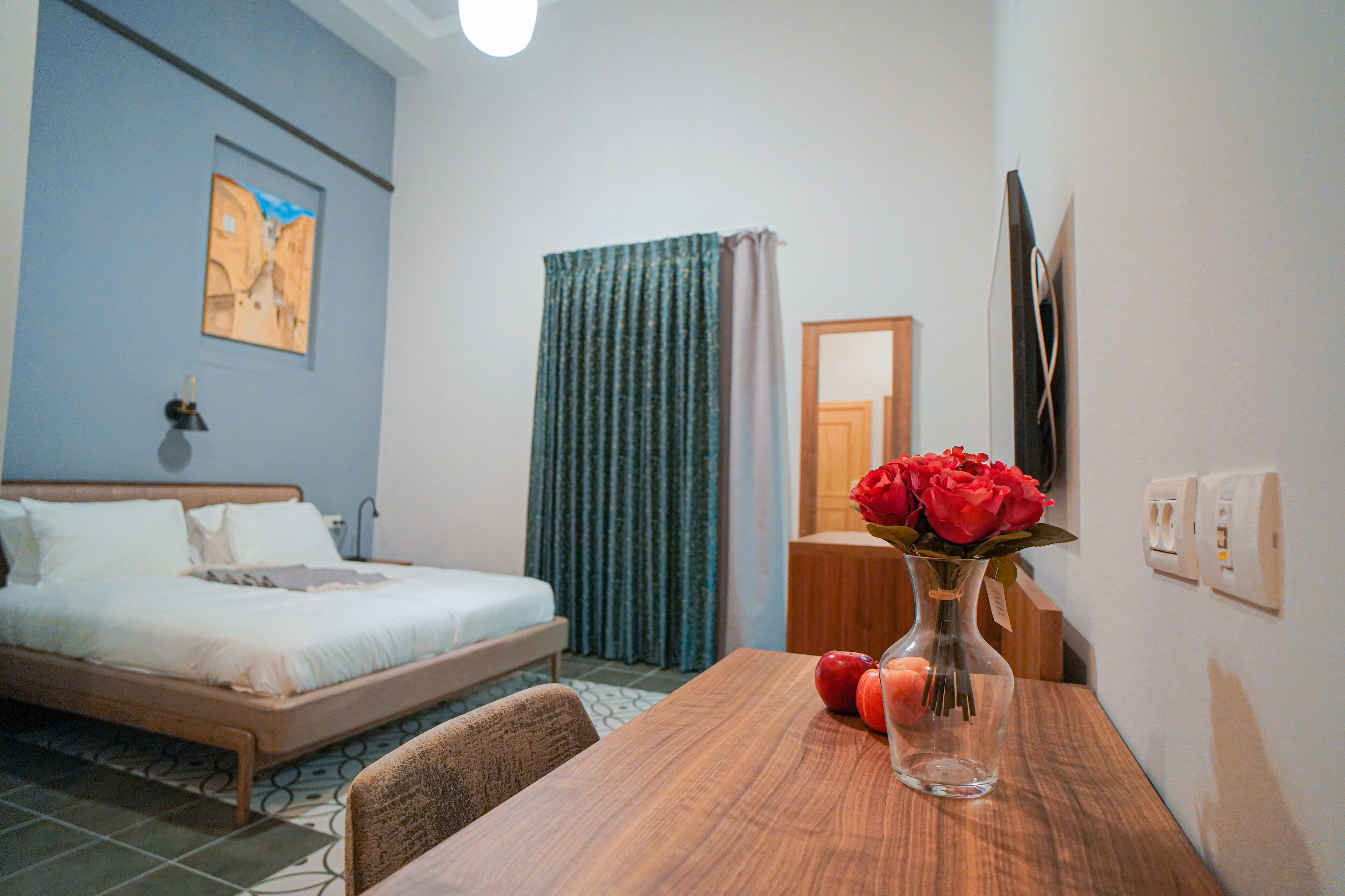 A hotel room with a bed, a wooden table, a vase with flowers, and a painting on the wall.