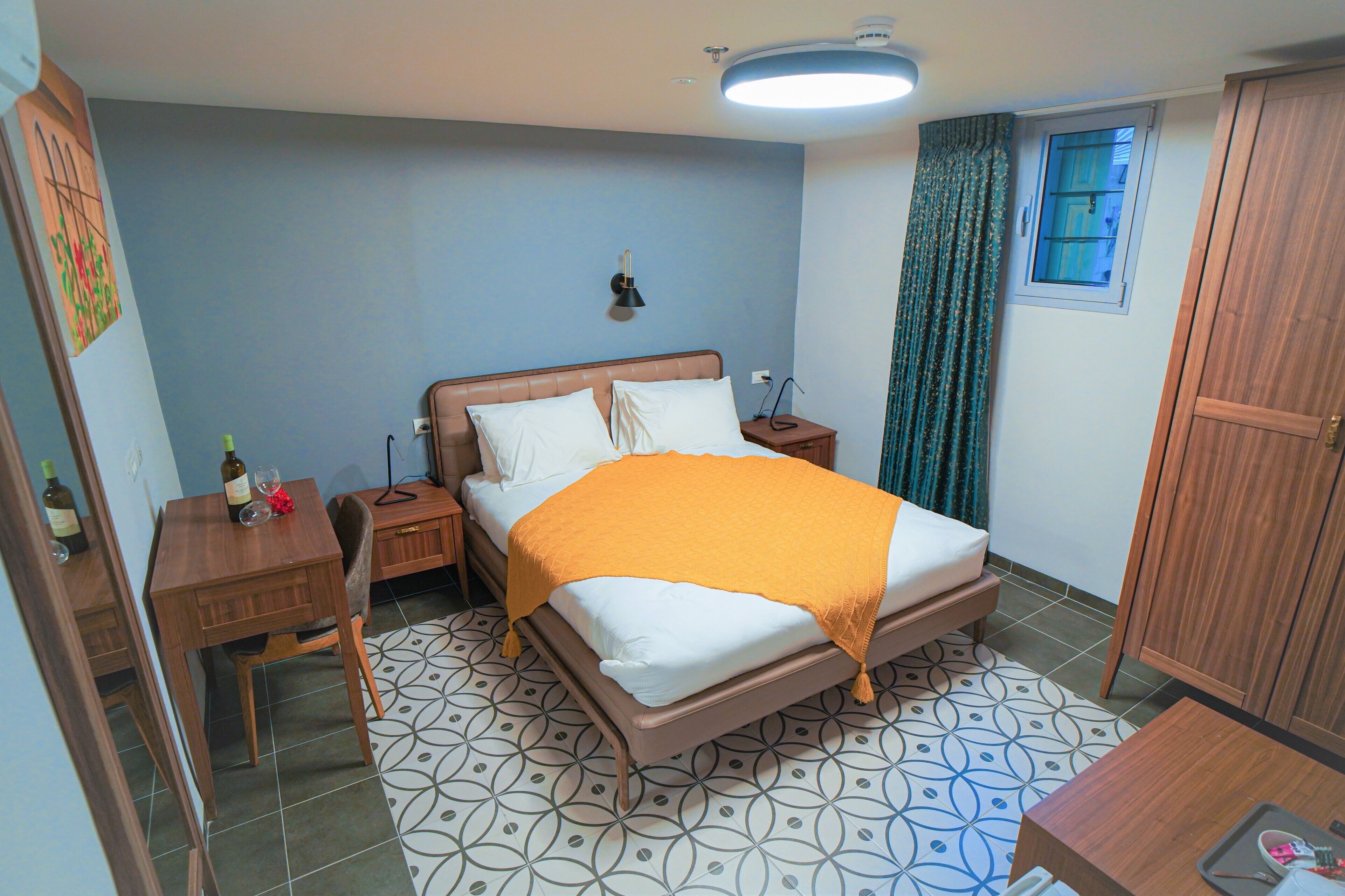 A bedroom with a bed, bedside tables, a nightstand, a wooden wardrobe, a window with curtains, and a patterned floor.