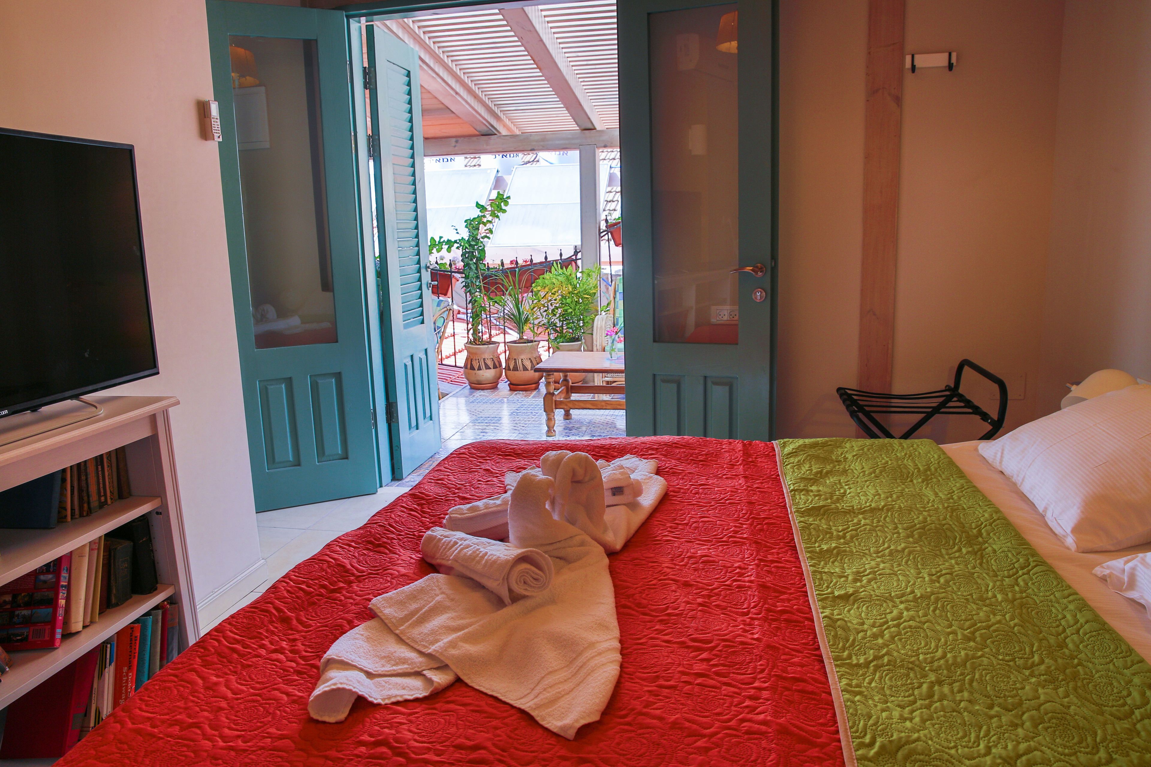 A hotel room with a red and green bedspread, a TV on the left, and a view of the outdoor area through open doors.