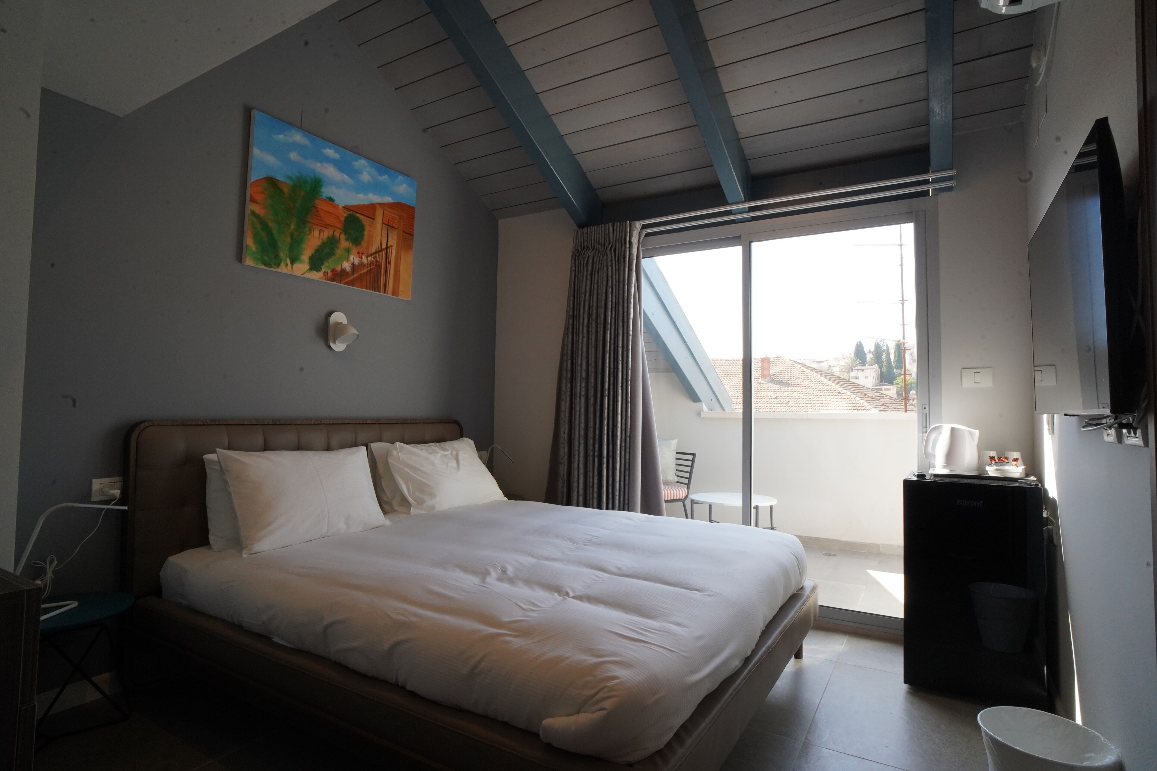 A hotel room with a bed, a painting, a desk, a chair, a television, and a balcony with a view.