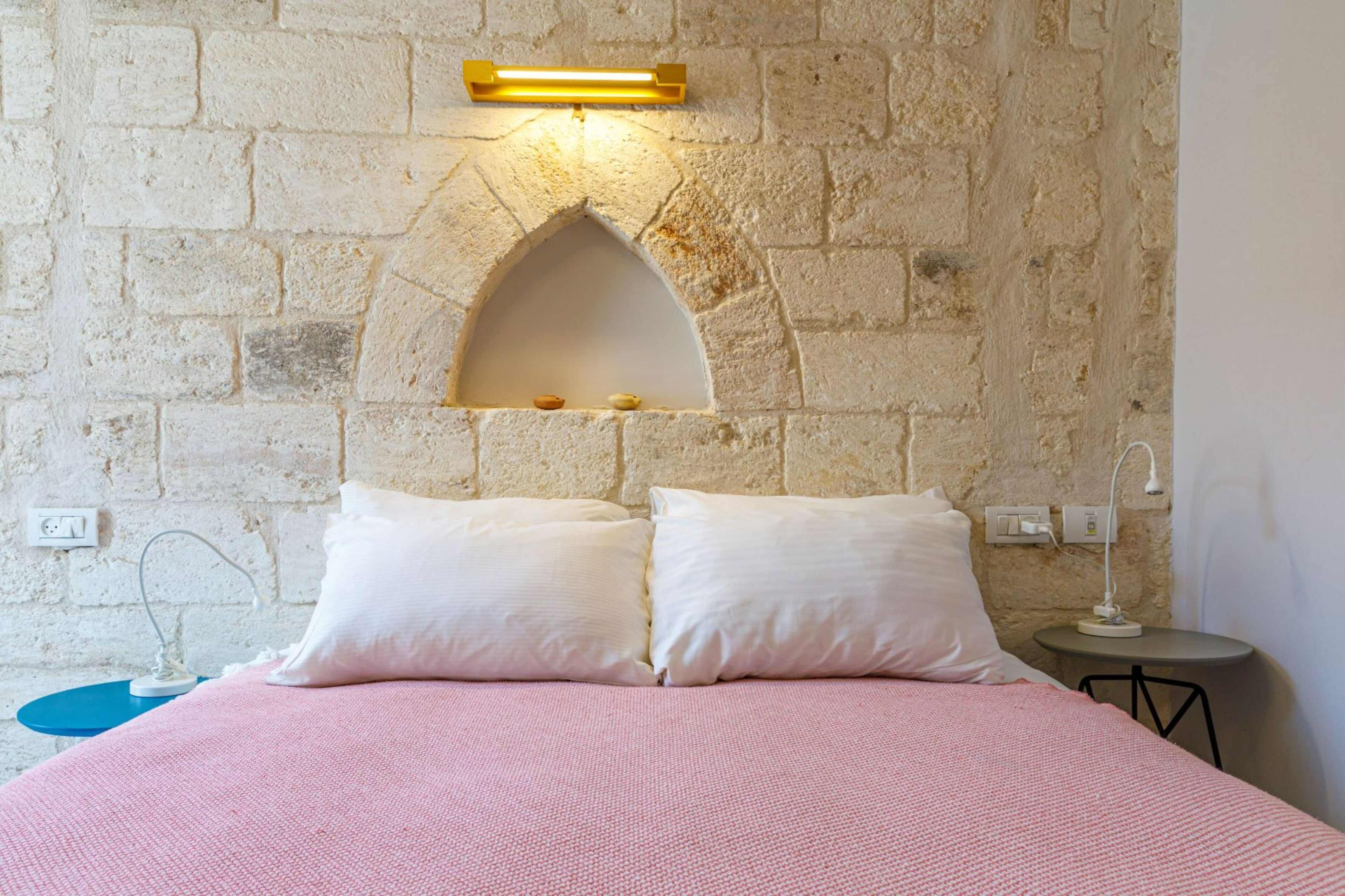 Un dormitorio con pared de piedra, una cama con ropa de cama rosa, dos almohadas blancas, una mesita con lámpara y un aplique de pared.