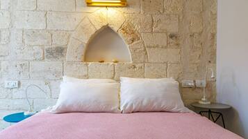 Un dormitorio con pared de piedra, una cama con ropa de cama rosa, dos almohadas blancas, una mesita con lámpara y un aplique de pared.