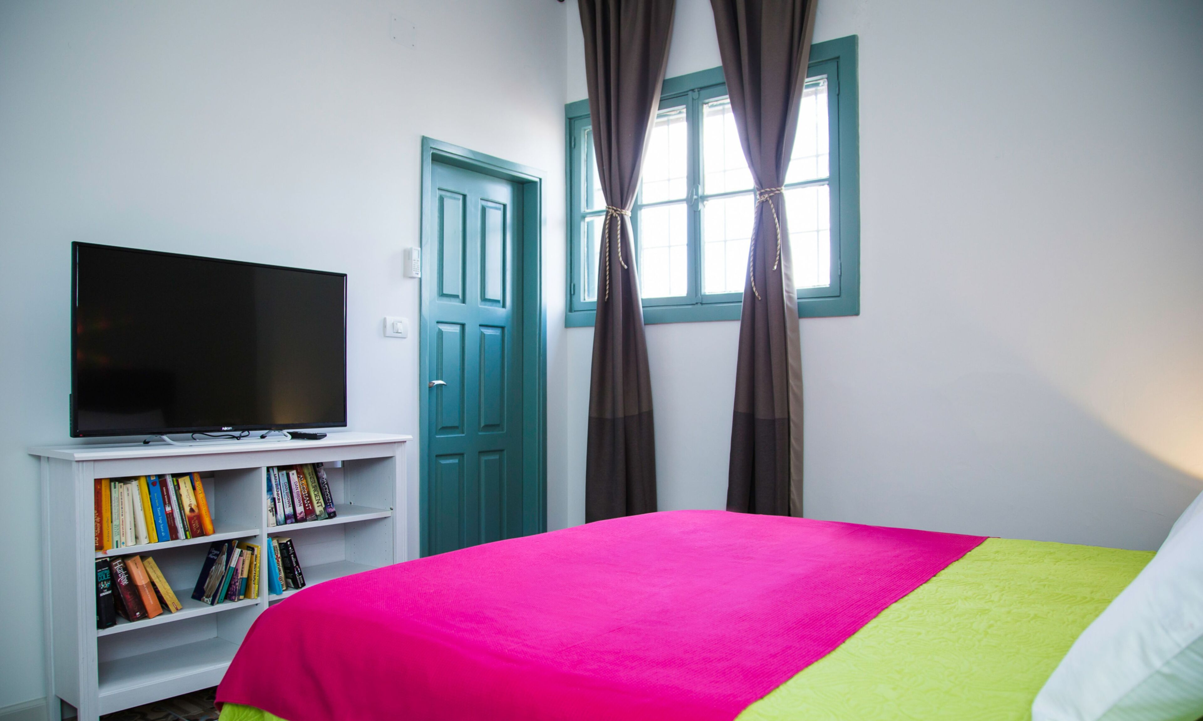 A bedroom with a bed, a television on a stand, a bookshelf, and a window with curtains.
