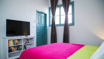 Un dormitorio con cama, televisión sobre un mueble bajo, estantería y ventana con cortinas.