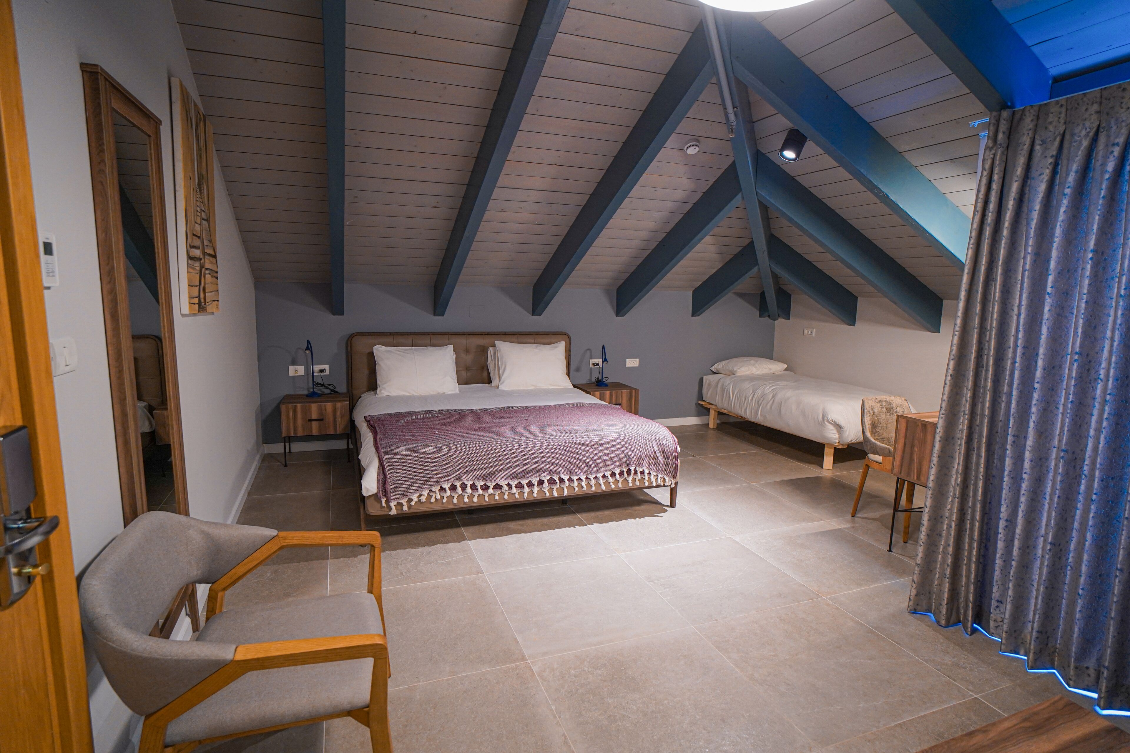 Un dormitorio con una cama grande, un escritorio y una silla. Hay vigas de madera en el techo y una ventana con cortinas.