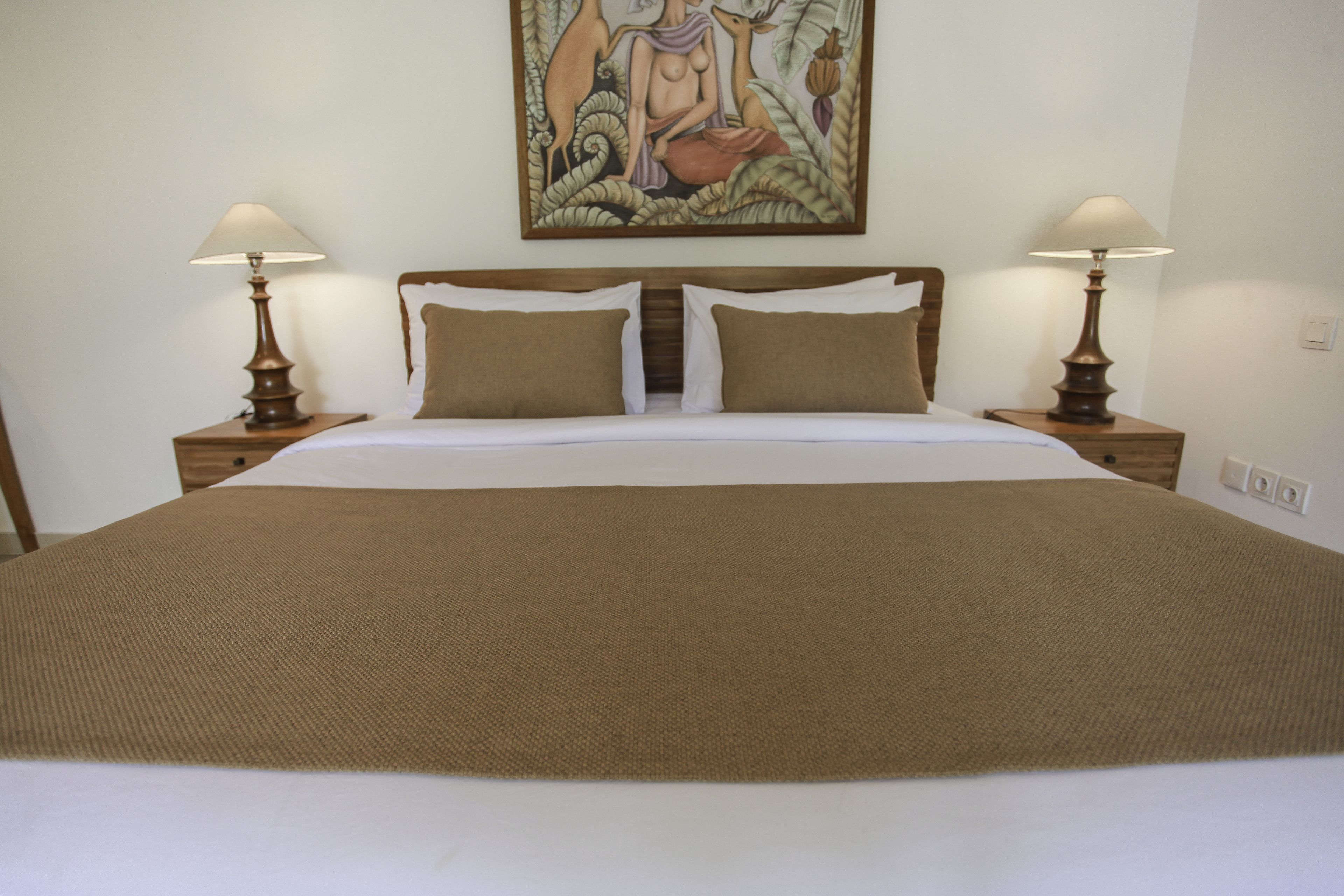 A large bed with a brown bedspread, two bedside tables with lamps, and a painting above the bed.