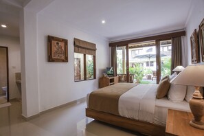 A bedroom with a bed, a TV, a lamp, and a window with a view of the outside. - Jati Cottage (Ubud)