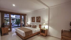 A modern hotel room with a large bed, two bedside tables with lamps, a wooden chair, and a view of the outdoors through large windows. - Jati Cottage (Ubud)