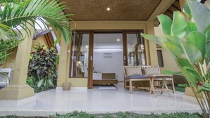 A well-maintained outdoor seating area with a wooden bench and a small table, surrounded by lush greenery and a clear pathway leading to a building entrance. - Jati Cottage (Ubud)