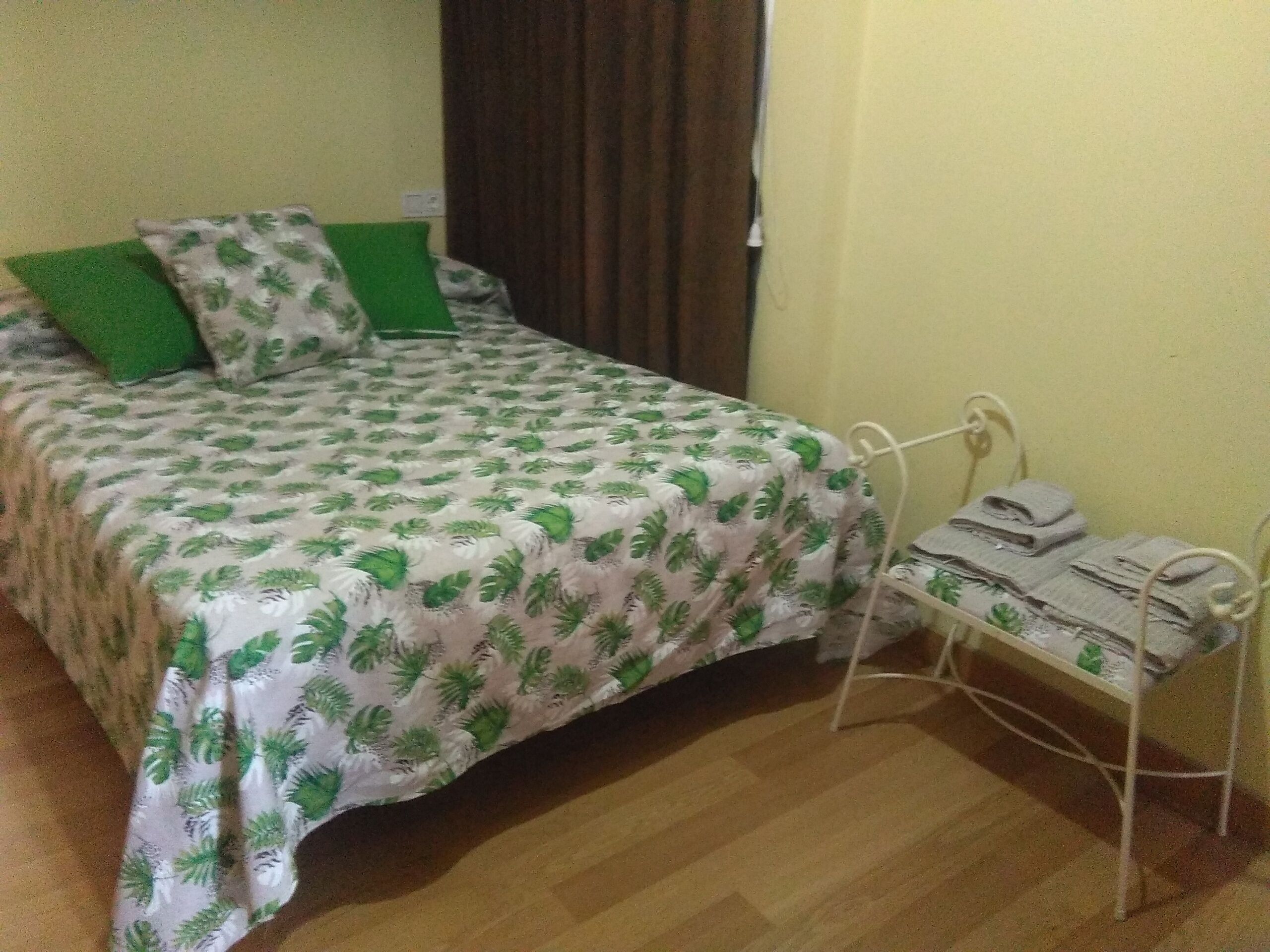 A bed with a green and white leaf-patterned bedspread, a green pillow, and a metal bedside table with a folded towel.