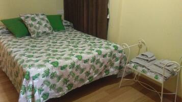 A bed with a green and white leaf-patterned bedspread, a green pillow, and a metal bedside table with a folded towel.