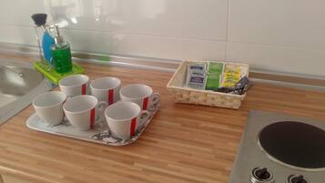 Una encimera de cocina con una bandeja de tazas blancas con detalles rojos, un dispensador de jabón verde y una cesta de mimbre con alimentos.