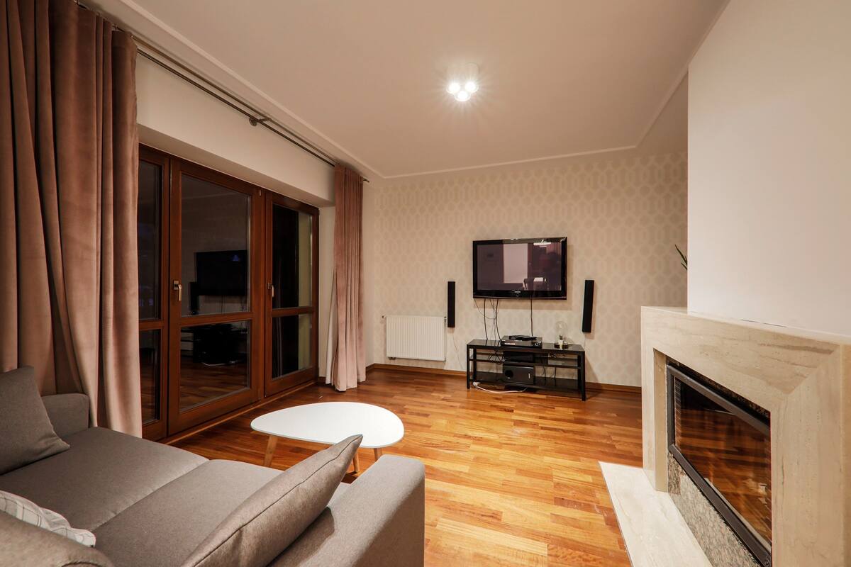 A living room with a sofa, a white coffee table, a fireplace, a TV mounted on the wall, and a wooden floor.