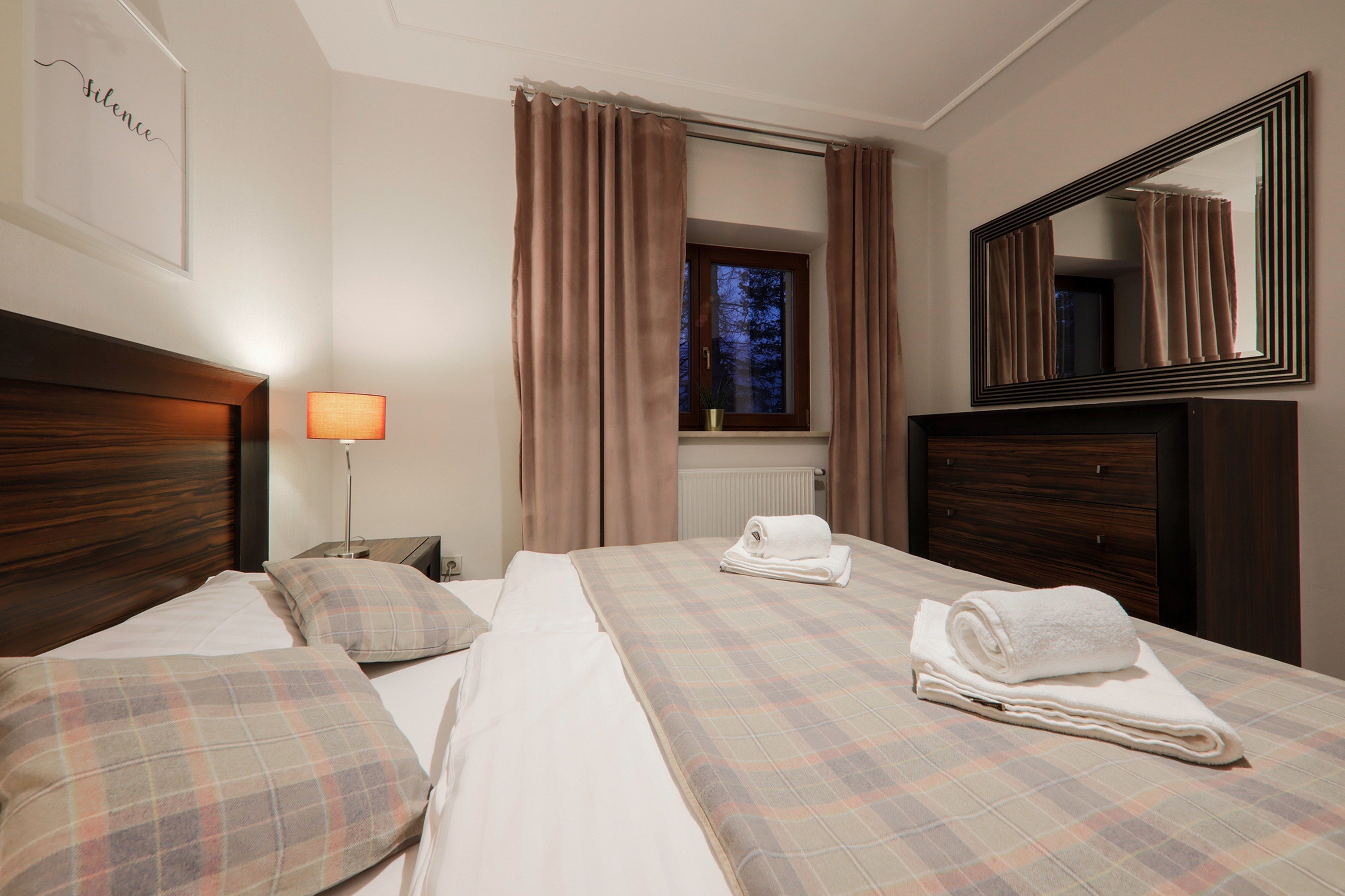 A hotel room with two beds, a window with curtains, a bedside lamp, a mirror, and a framed picture on the wall.