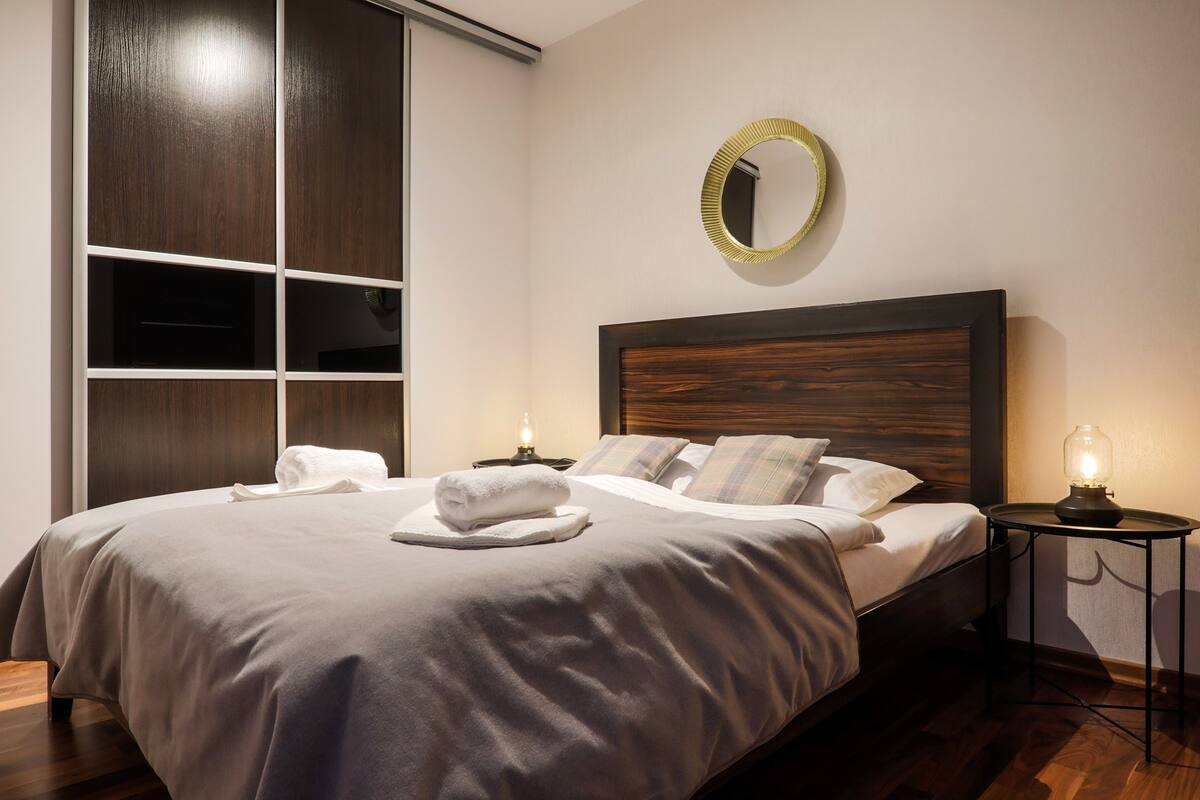 A neatly made bed with a white comforter and pillows, a bedside table with a lamp, and a large mirror on the wall.