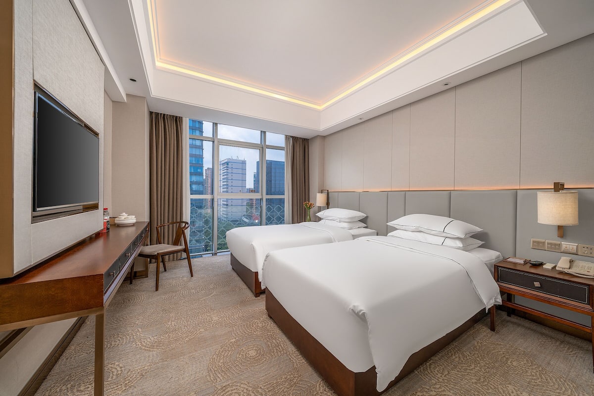 A hotel room with two beds, a desk, a TV, and a large window with a city view.