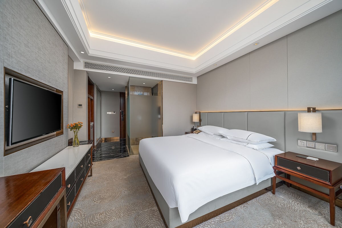 A hotel room with a large bed, a TV mounted on the wall, a bedside table with a lamp, and a carpeted floor.