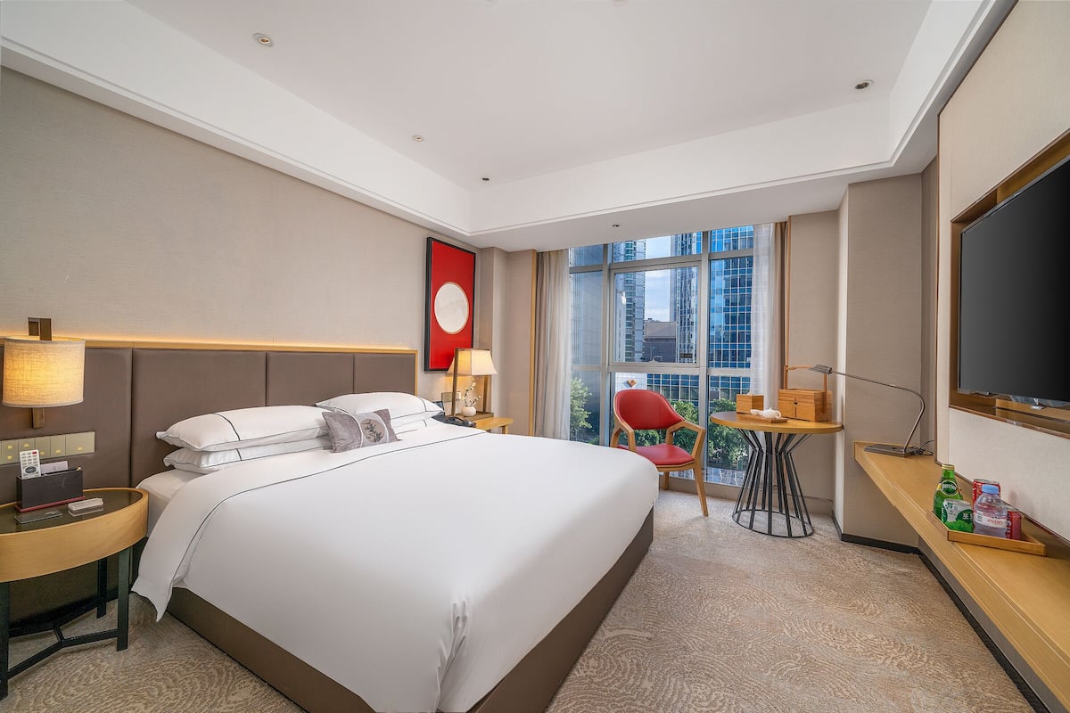 A hotel room with a large bed, a desk with a chair, a television, and a window with a city view.