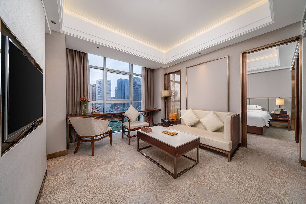 A hotel room with a large window, a flat-screen TV, a sofa, a coffee table, and a dining area with a table and chairs.