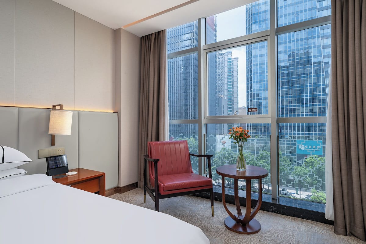A hotel room with a bed, a desk with a lamp, a chair, a small table with a vase, and a large window with a city view.