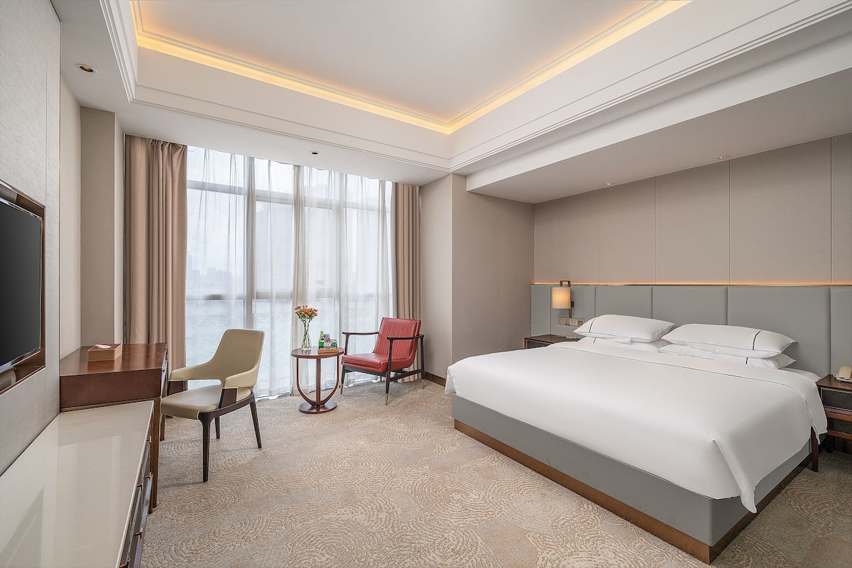 A hotel room with a large bed, a desk, a chair, a small table with a vase, and a window with curtains.