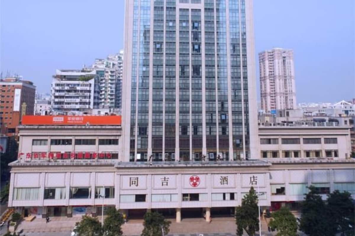 A multi-story building with a prominent glass facade, a red sign on the ground floor, and several smaller buildings in the background.