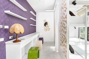 A modern bathroom with purple walls, white shelves, a green stool, a yellow lamp, and a tree decal on the wall.
