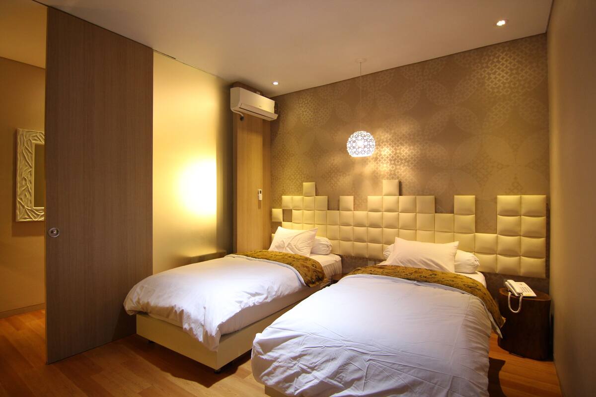 A hotel room with two beds, a wall-mounted air conditioner, a decorative light fixture, and a bedside telephone.