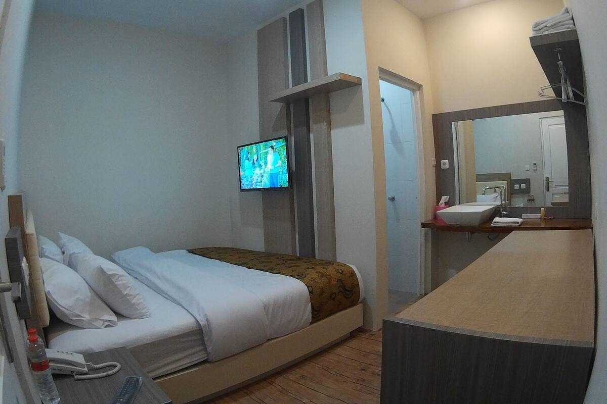 A hotel room with a bed, a TV mounted on the wall, a bathroom with a sink and mirror, and a wooden floor.