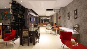 Un hall d’hôtel comprenant un comptoir d’accueil, des fauteuils rouges, un espace repas avec des tables et un mur orné de motifs.