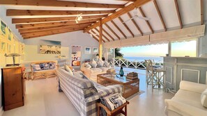 A spacious living room with a high ceiling, wooden beams, a large sofa set, a coffee table, and a view of the sea. - Cape Rose Cottage (Cape Town)