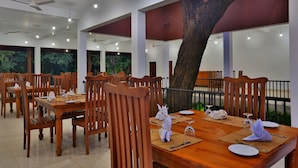 Un restaurant avec des tables et des chaises en bois, une grande fresque représentant un arbre sur le mur et de grandes fenêtres donnant sur un paysage verdoyant.
