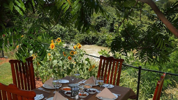 Une table à manger installée à l’extérieur, avec des assiettes, des verres et des couverts, entourée de chaises et recouverte d’une nappe, et offrant une vue sur une rivière et la verdure en arrière-plan.
