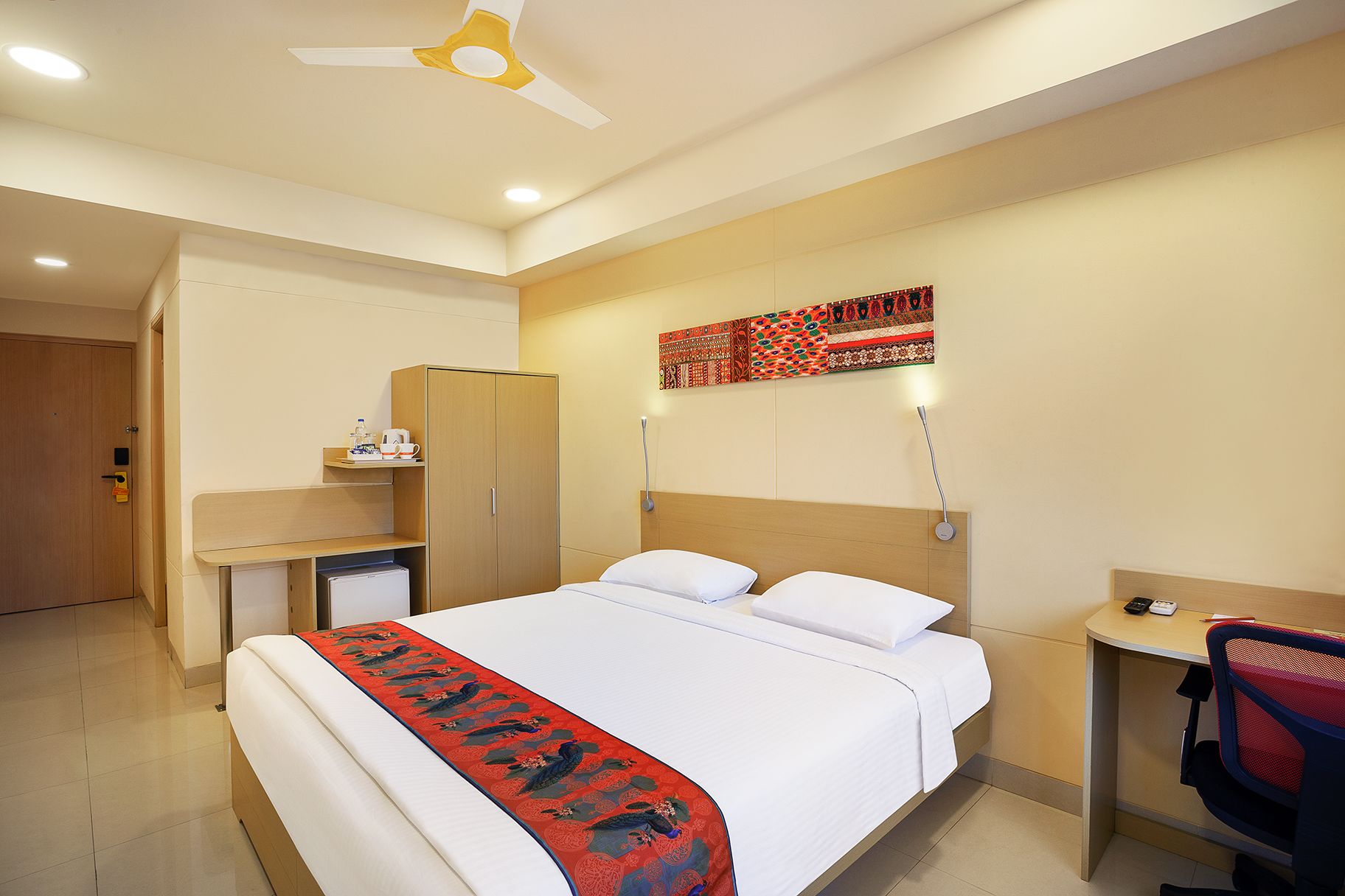 Habitación de hotel con cama, escritorio, armario, ventilador de techo y una obra de arte colgada en la pared.