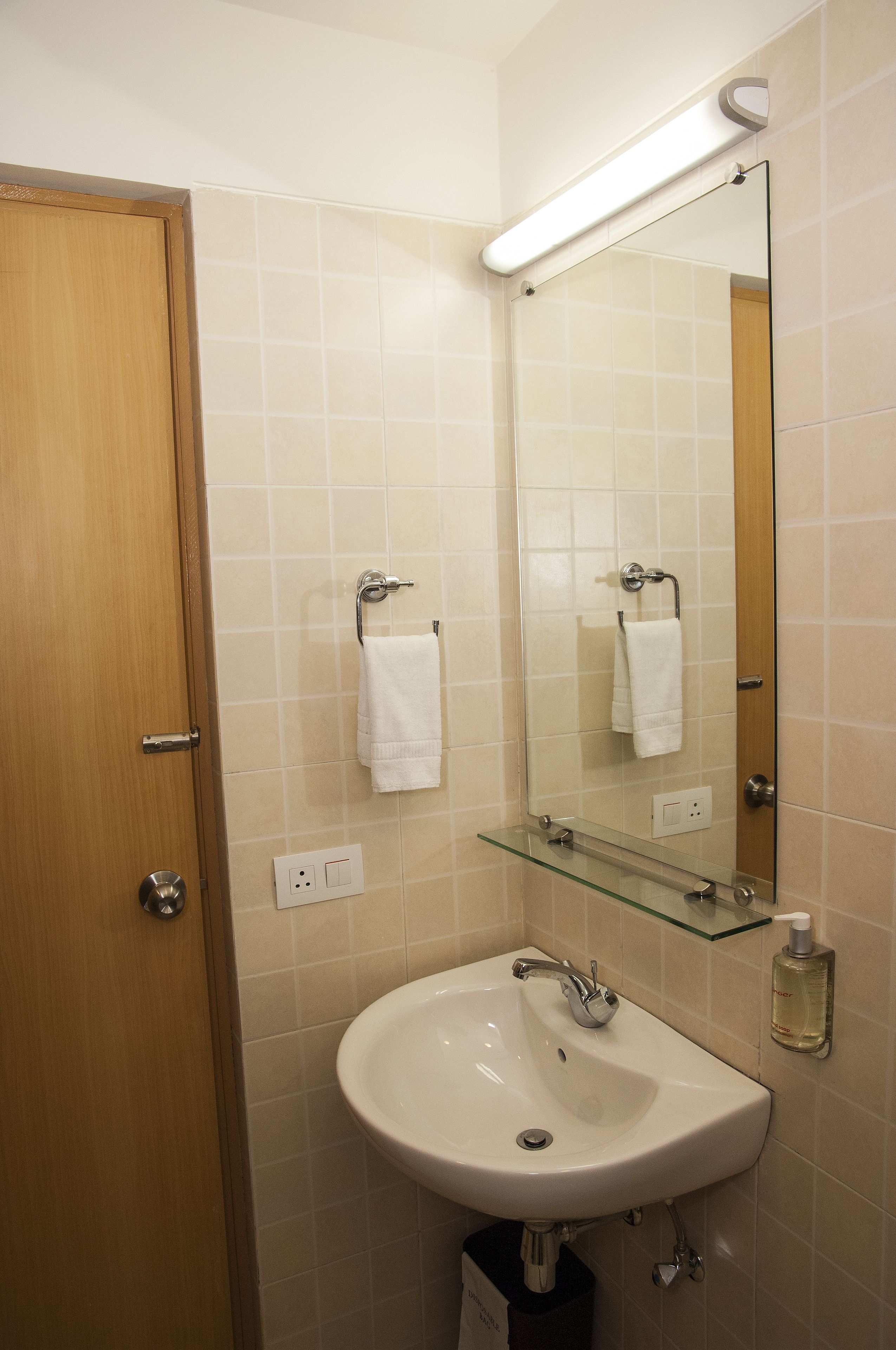 A bathroom with a wooden door, a white sink with a glass shelf above it, a mirror, and a towel hanging on the wall.