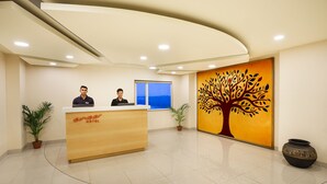 A hotel reception area with a wooden reception desk, two receptionists, a large tree mural, and a window with a view. - Ginger Vapi (Vapi)