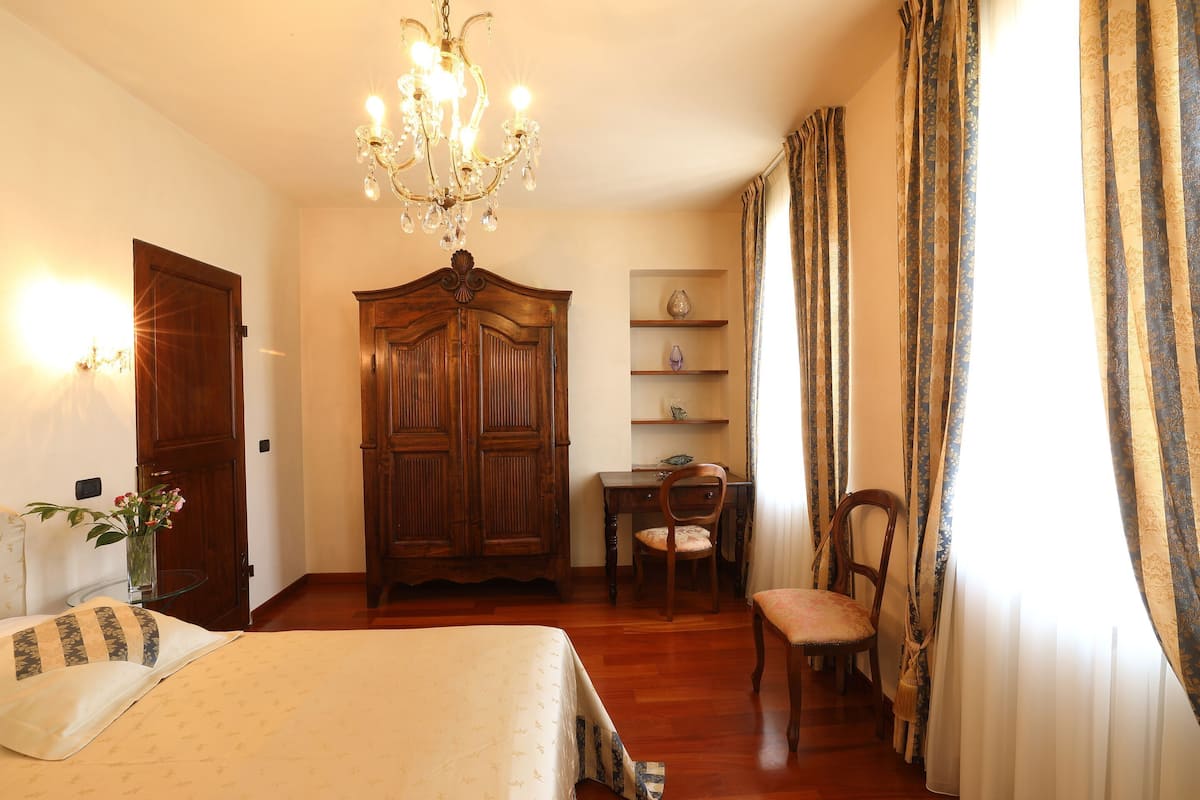 A bedroom with a bed, a wooden wardrobe, a desk with a chair, a chandelier, and curtains.