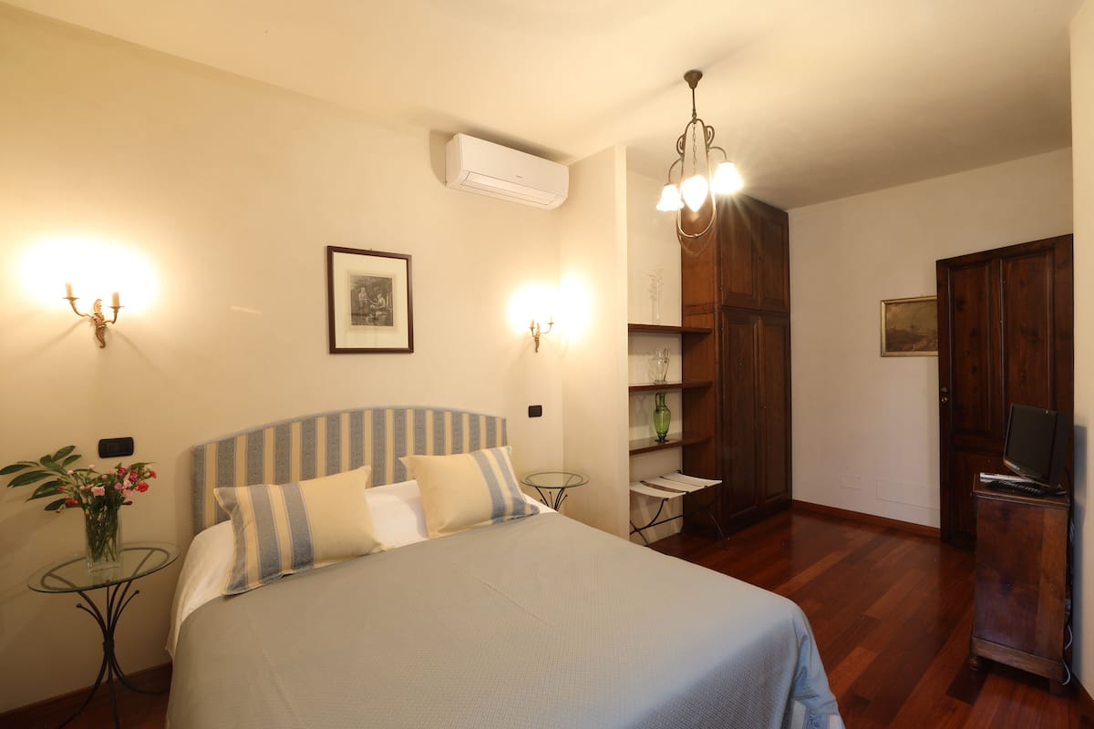A bedroom with a bed, bedside tables, a small table with a vase of flowers, a wall-mounted air conditioner, a chandelier, and a television on a wooden cabinet.
